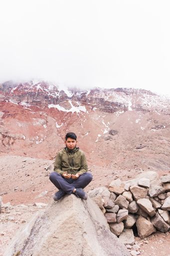A vertical shot of the young man in the mountains, sitting on the rock and meditating.