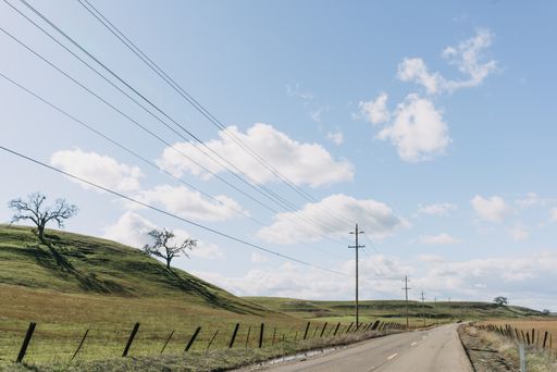 A wide shot of a highway road near green hills under a clear blue sky with clouds