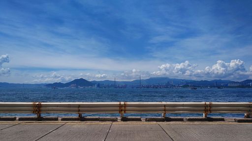 A scenic view of the ocean with distant mountains under a clear blue sky, featuring a rusted guardrail in the foreground.