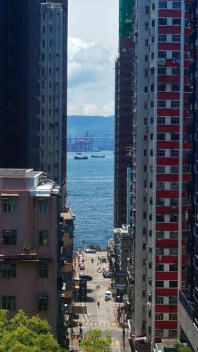 Urban view of a street leading to the sea between tall buildings in Hong Kong, with a clear sky and distant ships.