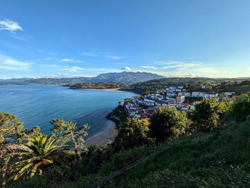 Scenic view of a coastal town with lush greenery, sandy beach, and mountains under a clear blue sky in Asturias, Spain.