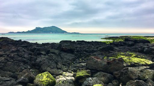 Scenic view of a rocky coastline with a distant island under a cloudy sky, showcasing natural beauty and tranquility.