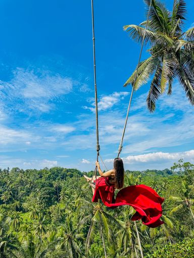 A woman in a flowing red dress swings over lush tropical forest under a vibrant blue sky in Bali, Indonesia.