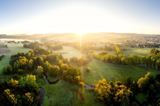 Aerial view of a lush green landscape at sunrise with a small town in the distance, showcasing vibrant foliage and a serene atmosphere.