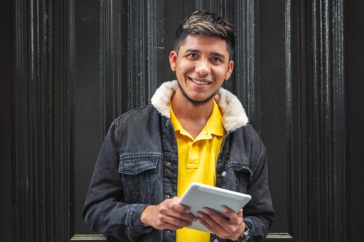 A young man with a yellow shirt looking at the camera and smiling holding a tablet with a black background.