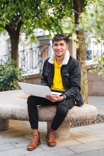 A vertical shot of a young man sitting on a stone bench in the street with a laptop looking ahead and smiling.