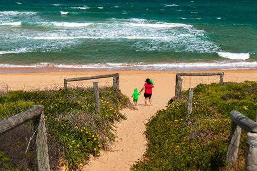 A mother and two children walk towards a sandy beach with vibrant blue waves under a clear sky, surrounded by greenery and wooden fences.