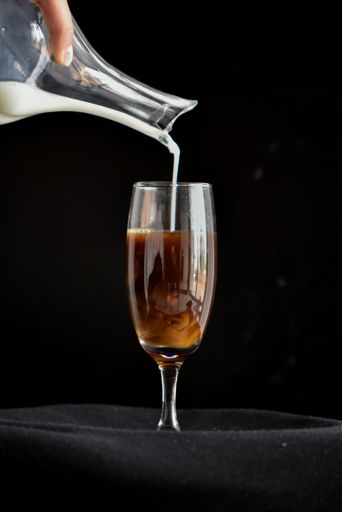 A vertical shot of a man pouring milk inside a champagne flute glass full of coffee; black background