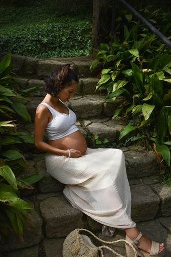 A vertical shot of a Caucasian, pregnant woman in white summer clothes sitting in apark, looking at her belly