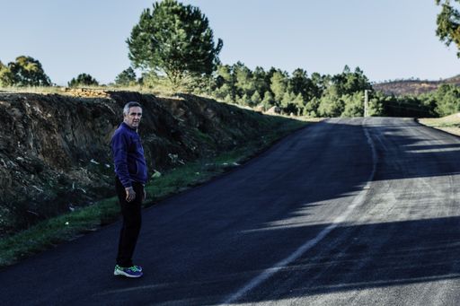 An old man in a blue jacket standing on the road surrounded by rocks and greenery under the sunlight