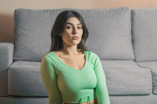 A young woman with long, dark hair is seated in front of a gray sofa looks directly at the camera