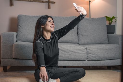 Young woman in sportswear taking a selfie while stretching in a cozy living room setting with a gray sofa and warm lighting.