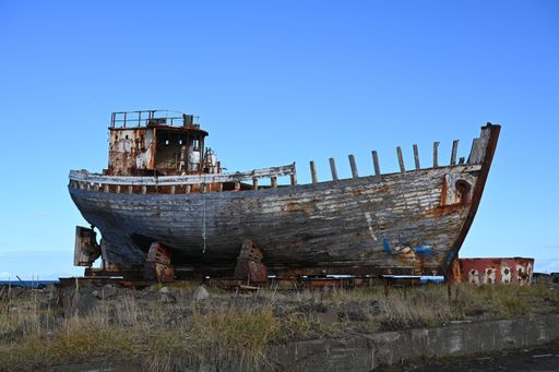 An old, rusted fishing boat sits abandoned on a grassy shore under a clear blue sky.