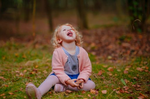 A shallow focus shot of a happy little girl sitting on the green grass in the park with her face up, an open mouth and stretched tongue