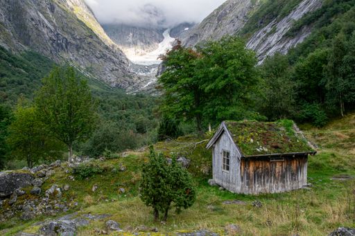 A rustic cabin with a green roof nestled in a lush valley surrounded by majestic mountains under a cloudy sky.