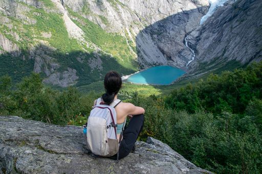 A woman with a backpack sits on a rock, overlooking a turquoise lake and lush valley in a mountainous landscape.