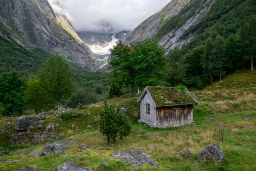 A rustic cabin with a moss-covered roof set in a lush green valley surrounded by towering mountains and a cloudy sky, creating a serene landscape.