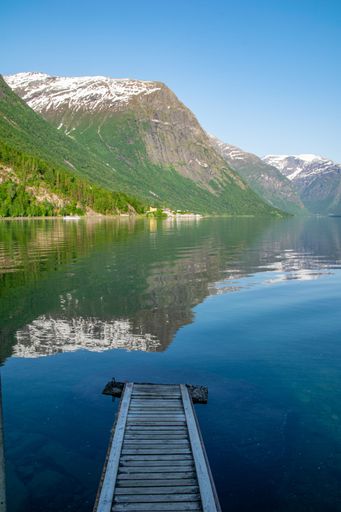 Scenic view of a wooden dock leading to a tranquil lake with snow-capped mountains and clear blue skies in the background.