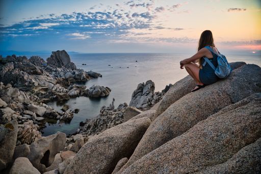 A closeup shot of a young woman with a backpack sitting on the stone looking to sunset at Capo Testa Santa, Italy