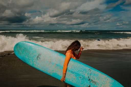 A woman with long hair carries a blue surfboard on a sandy beach with waves crashing under a cloudy sky.