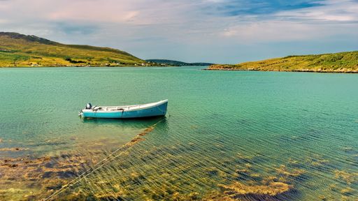 A sailboat is seen sailing in the middle of a tranquil body of water, surrounded by lush green hills and land in the backdrop