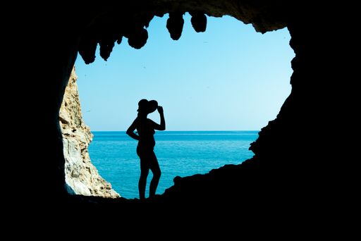 A silhouette of a female with a hat standing at the entrance of a rocky cave over sunny blue water