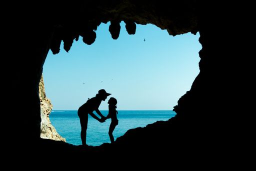A silhouette of a female with a kid standing at the entrance of a rocky cave over a sunny blue water