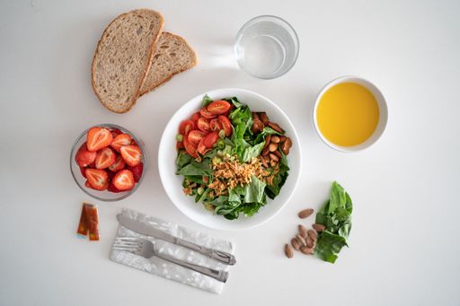 A top view of a bowl of salad with herbs, tomatoes, and almonds on the table with strawberries and bread slices