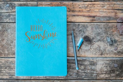 An overhead shot of a "hello sunshine" blue textbook with a ball-point pen next to it on wooden surface