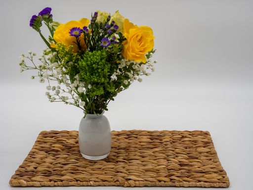 A picture of multicolor flowers in a vase on a straw tray against a grey background