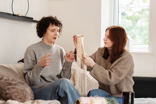 A curly Caucasian female knitting and a redhead female sitting next to her and helping