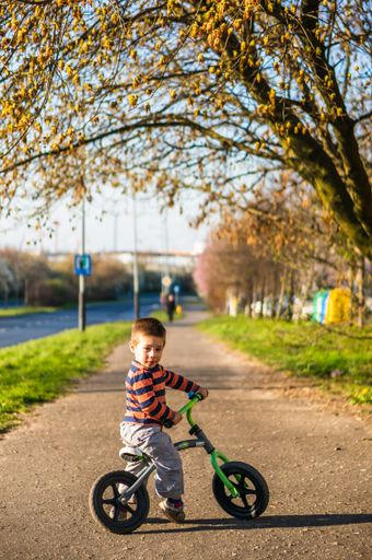 Small two years old Polish Caucasian boy sitting on a running bike on a footpath along grass and trees.
