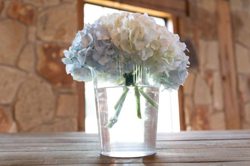 A close-up of white and blue hydrangeas in a clear vase on a wooden table with a stone wall background.