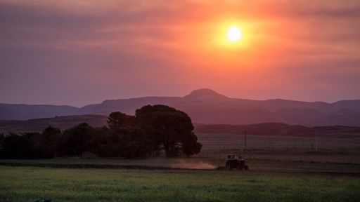A tractor plows a field under a vibrant sunset with mountains in the background, creating a serene rural landscape.