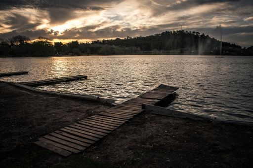 A serene lakeside scene at sunset with a wooden pier extending into the water, silhouetted trees, and a dramatic sky.
