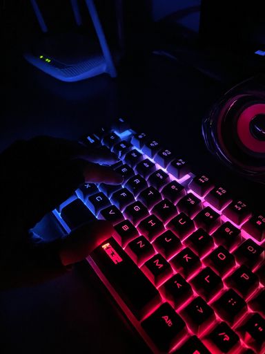 A vertical shot of a hand typing on an illuminated with colorful lights gaming  keyboard in a dark