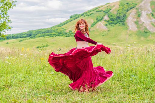 A young beautiful red-haired girl in a red ball gown is dancing a fiery dance on a green meadow against the backdrop of a relict mountain.