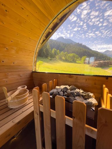 Interior of a wooden sauna with a panoramic window view of a lush green landscape and mountains under a partly cloudy sky.