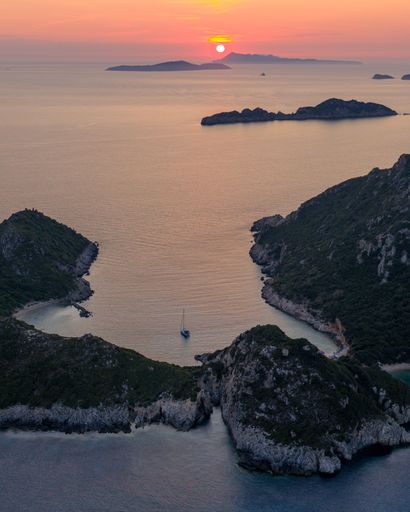 An aerial view of a serene bay with a sailboat and rocky coastline during a vibrant sunset in Corfu, Greece