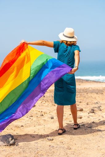A vertical shot of a lesbian person in a blue dress and white hat waving the LGBTQ flag by the sea