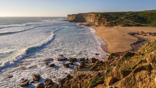 Scenic view of a sandy beach with rocky cliffs and waves under a clear sky at sunset.