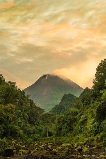 A stunning view of a volcano with lava flow surrounded by lush green forest under a dramatic sky.