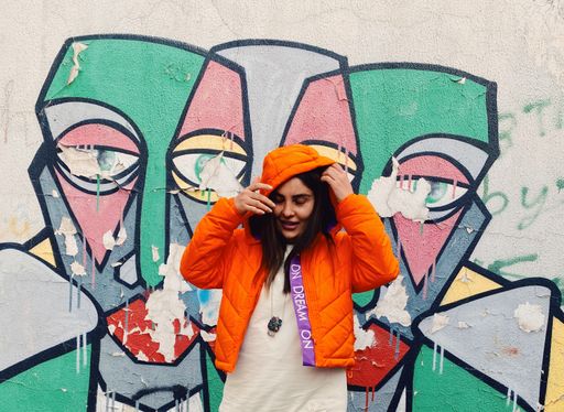A female in an orange jacket and a graffiti wall in the background.