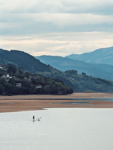 A serene view of a paddleboarder on a calm river with lush hills and distant mountains under a cloudy sky.