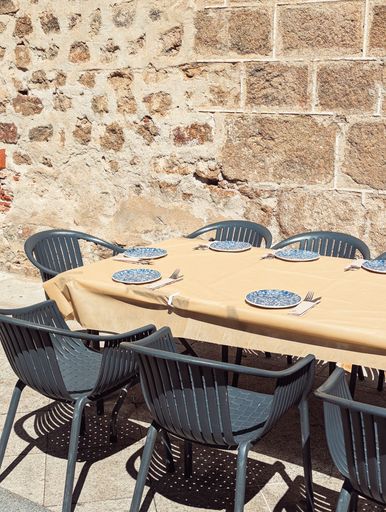 Outdoor dining setup with modern chairs and a table set against a rustic stone wall, under bright sunlight.