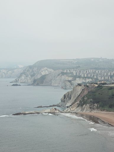 Coastal cliffs and buildings under a cloudy sky, overlooking the calm sea and sandy beach.