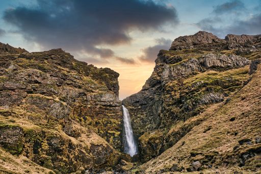 A waterfall flowing down the rocks covered with moss under a gray cloudy sky during the sunset