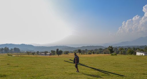 A male carrying two bamboos in a field surrounded by hills under the sunlight and a cloudy sky