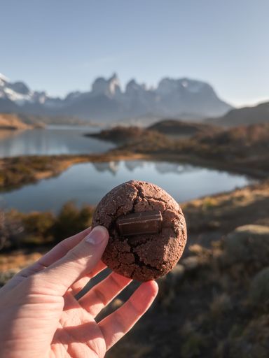 A hand holding a chocolate cookie with mountains and a lake in the background, capturing a scenic outdoor setting.