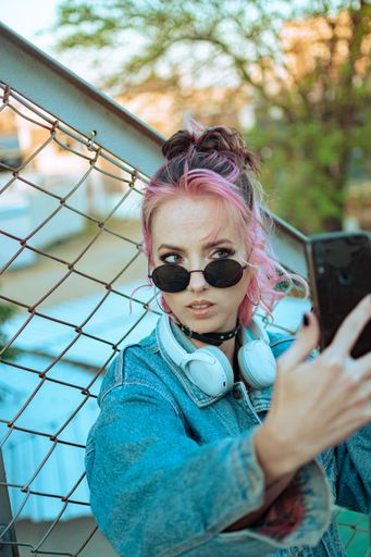 A vertical shot of a cool Caucasian lady with pink hair taking a selfie near a chain-link fence